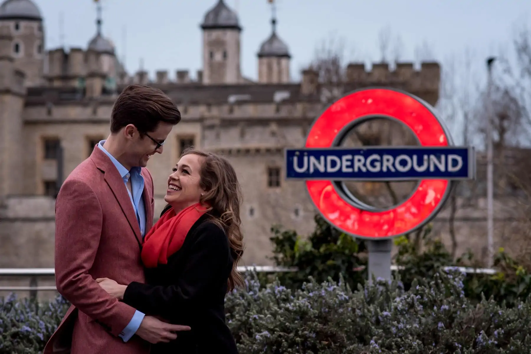 A couple laughing on their engagement shoot in from of the Tower of London and an Underground sign.