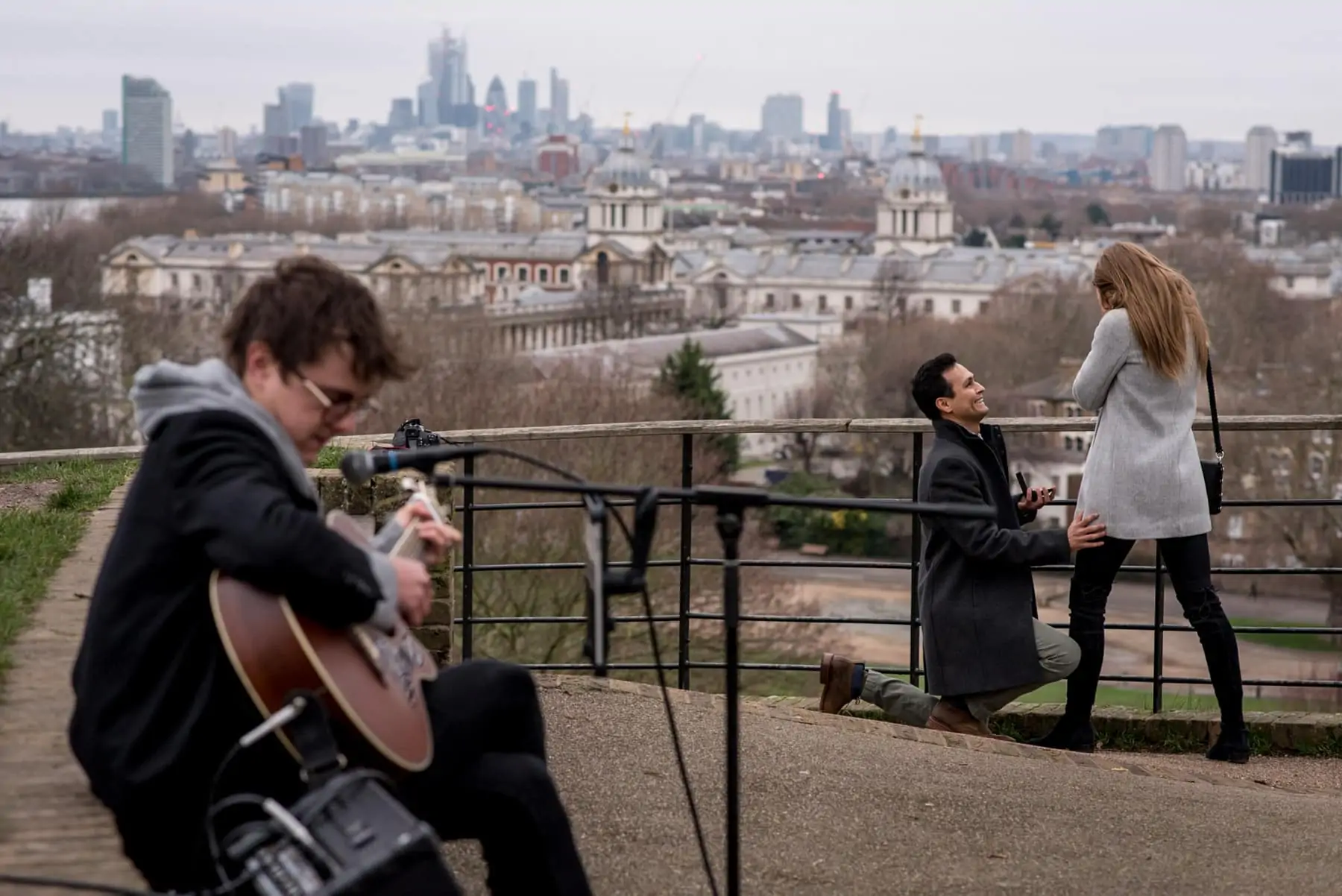 A man on his knees proposing to his girlfriend with views over London and a guitar player. Photo taken in Greenwich park.