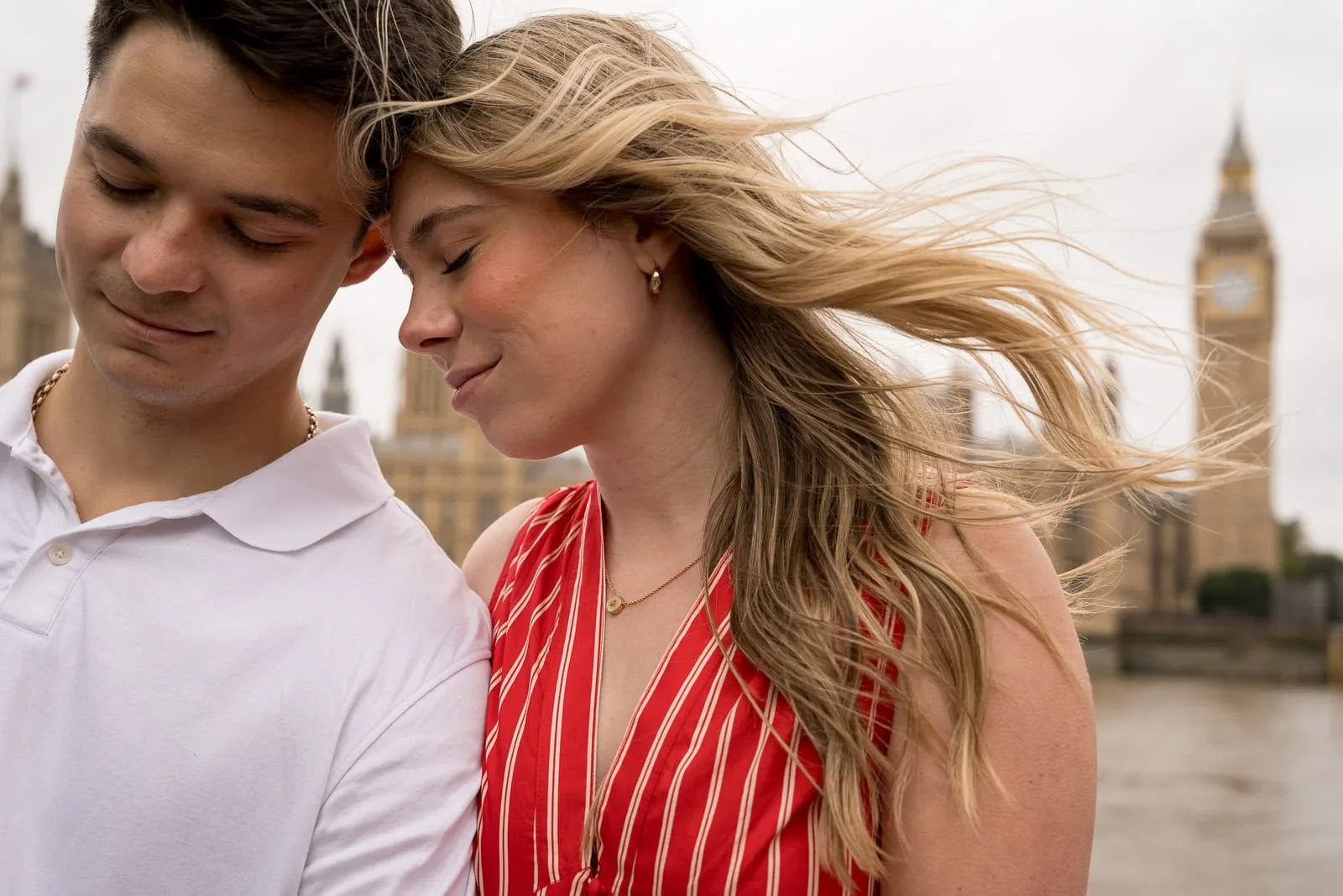 a couple touching heads with the girl in the red dress hair blond hair blowing in the wind with Big Ben in the background. They just got engaged!