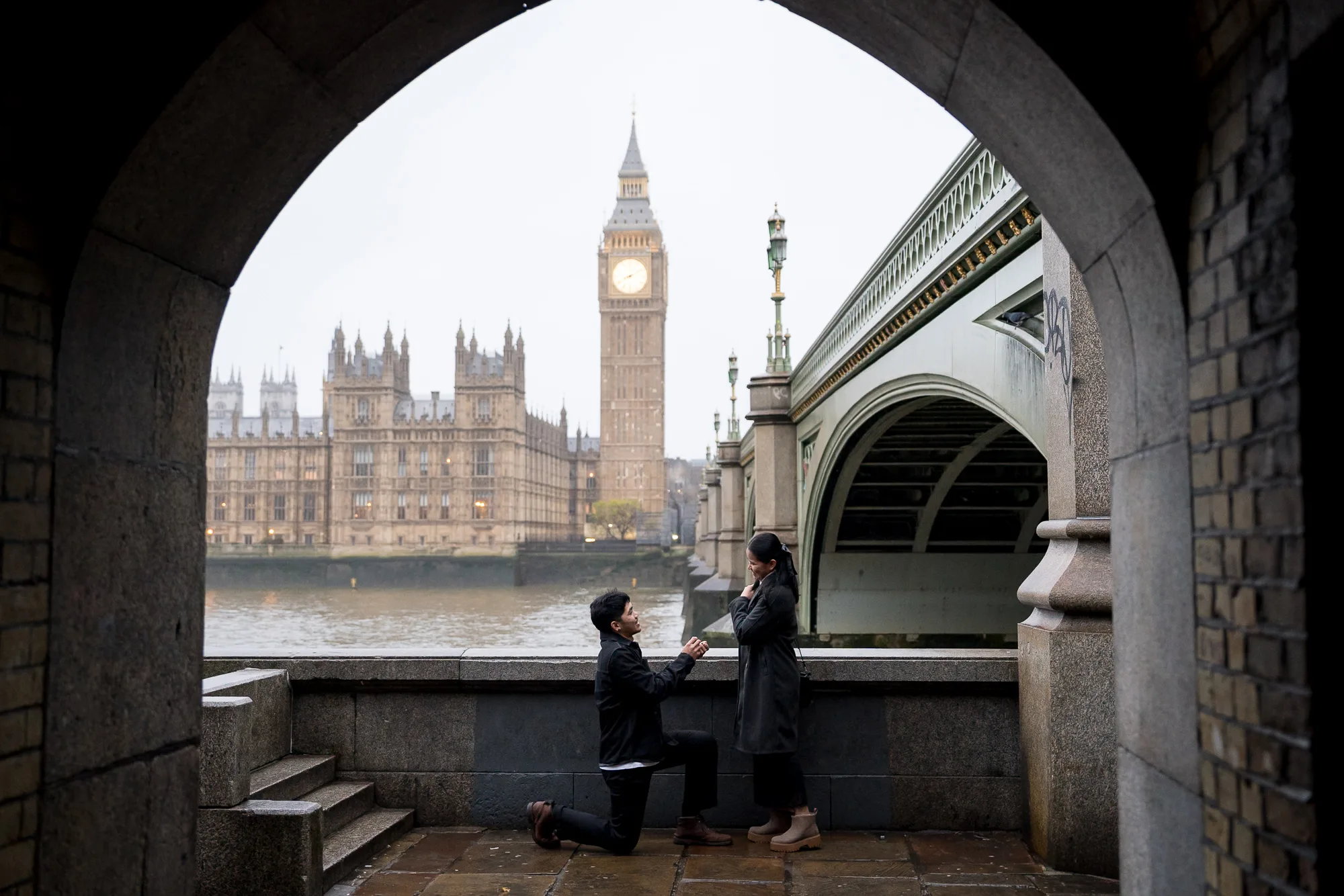 A man proposing to his girlfriend through an archway with Big Ben in the background, London