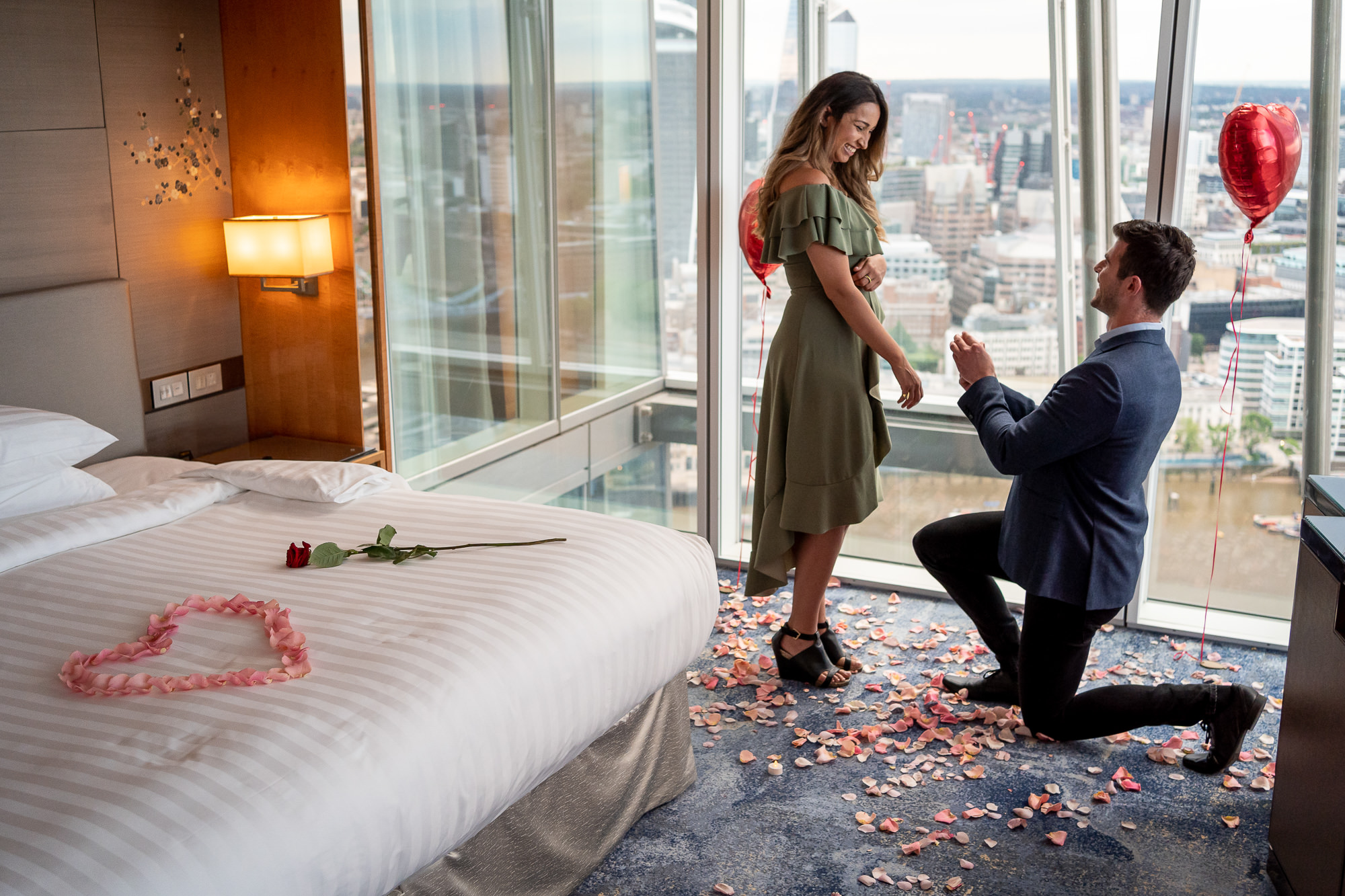 A man proposing to his girlfriend in their hotel room in the shard with red heart balloons, roses and petals.