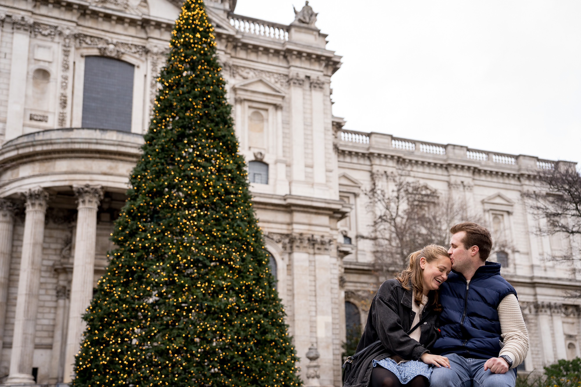 A couple kiss and laugh in front of the St Paul's Christmas Street during their proposal photo shoot