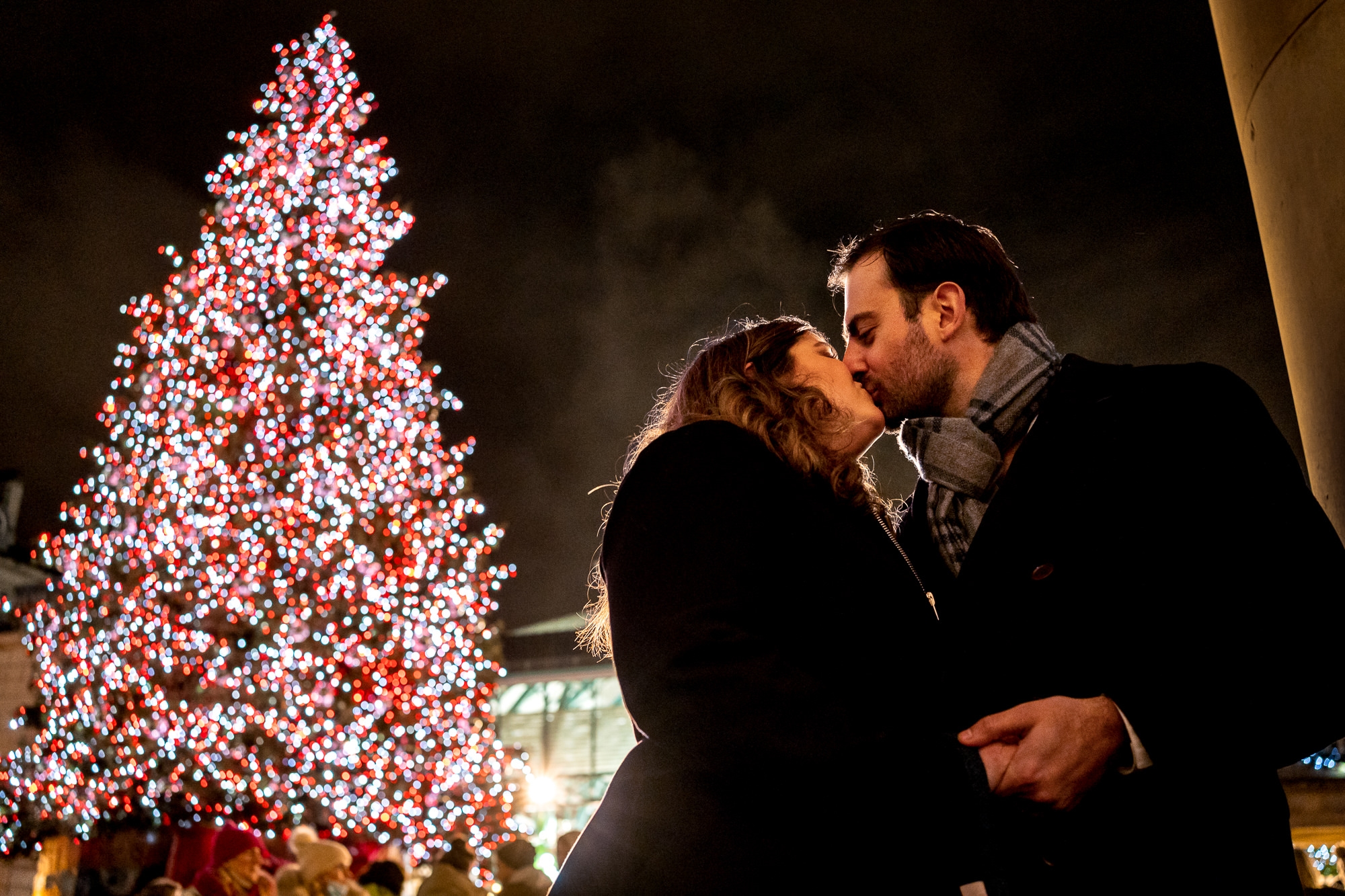 A couple kisses in front of the Covent Garden Christmas tree at night after she said yes to his marriage proposal