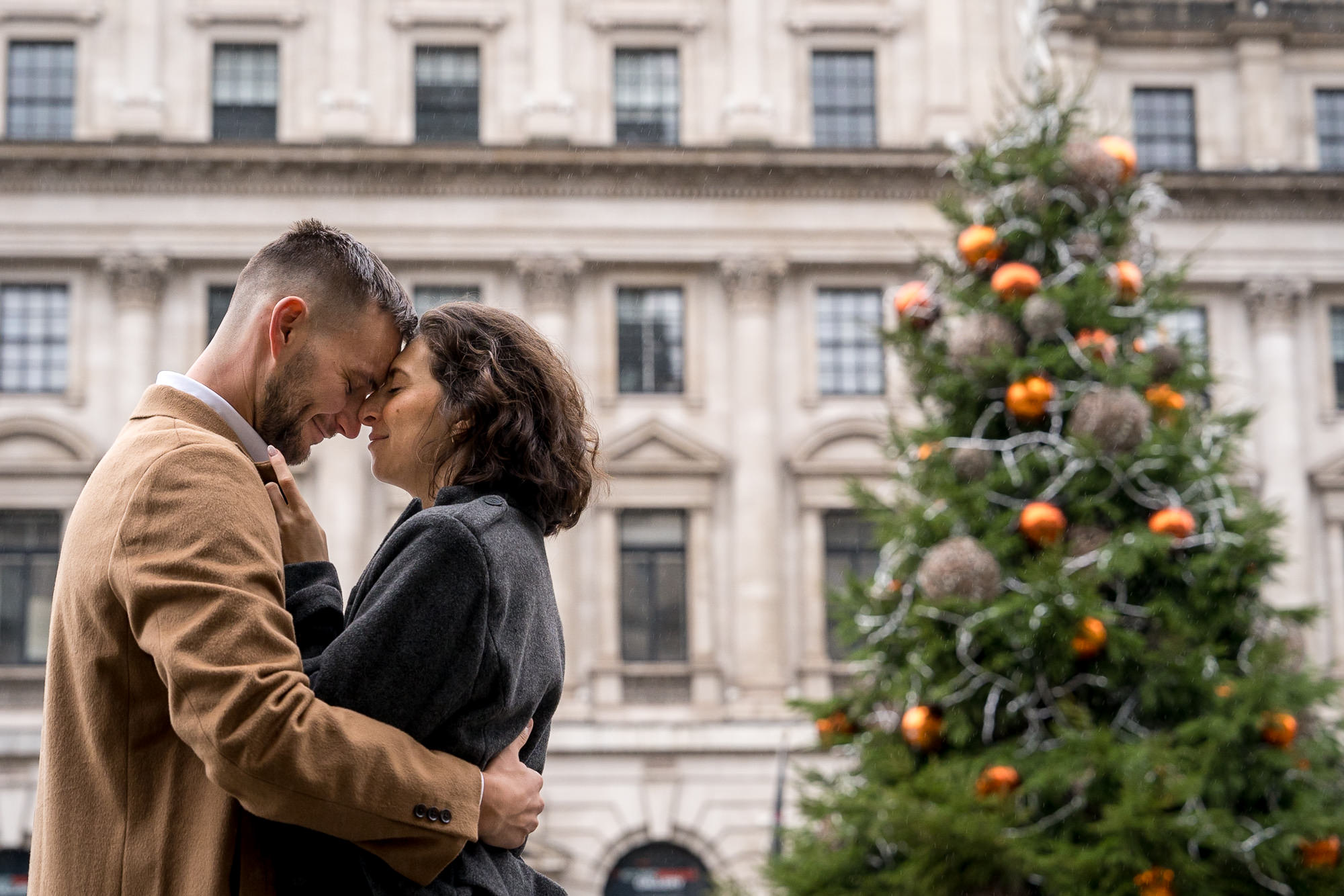 A couple touch heads after proposing in front of a Christmas tree in London