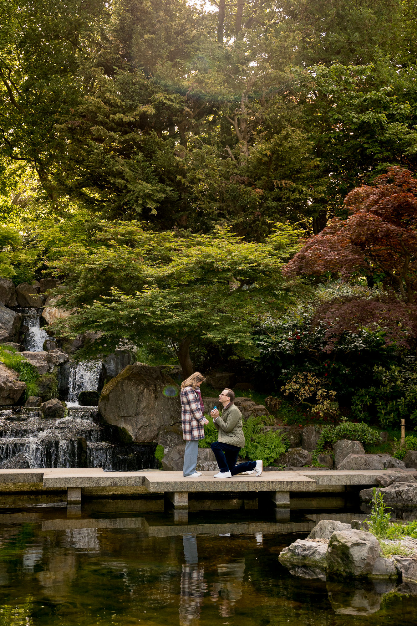 a man on one knee proposing to his girlfriend on the foot bridge in Kyoto Garden with the waterfall behind them. She said yes