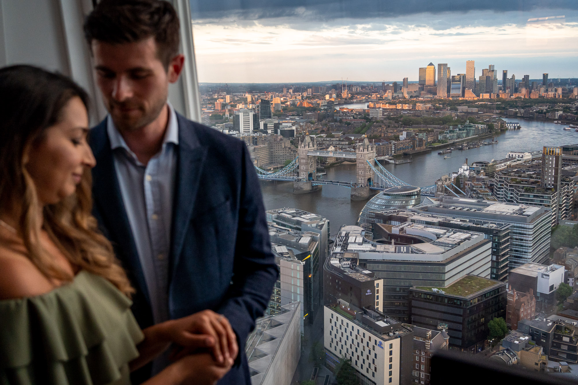 A couple standing by the window of their hotel room in the Shard looking out over London at sunset, following their proposal