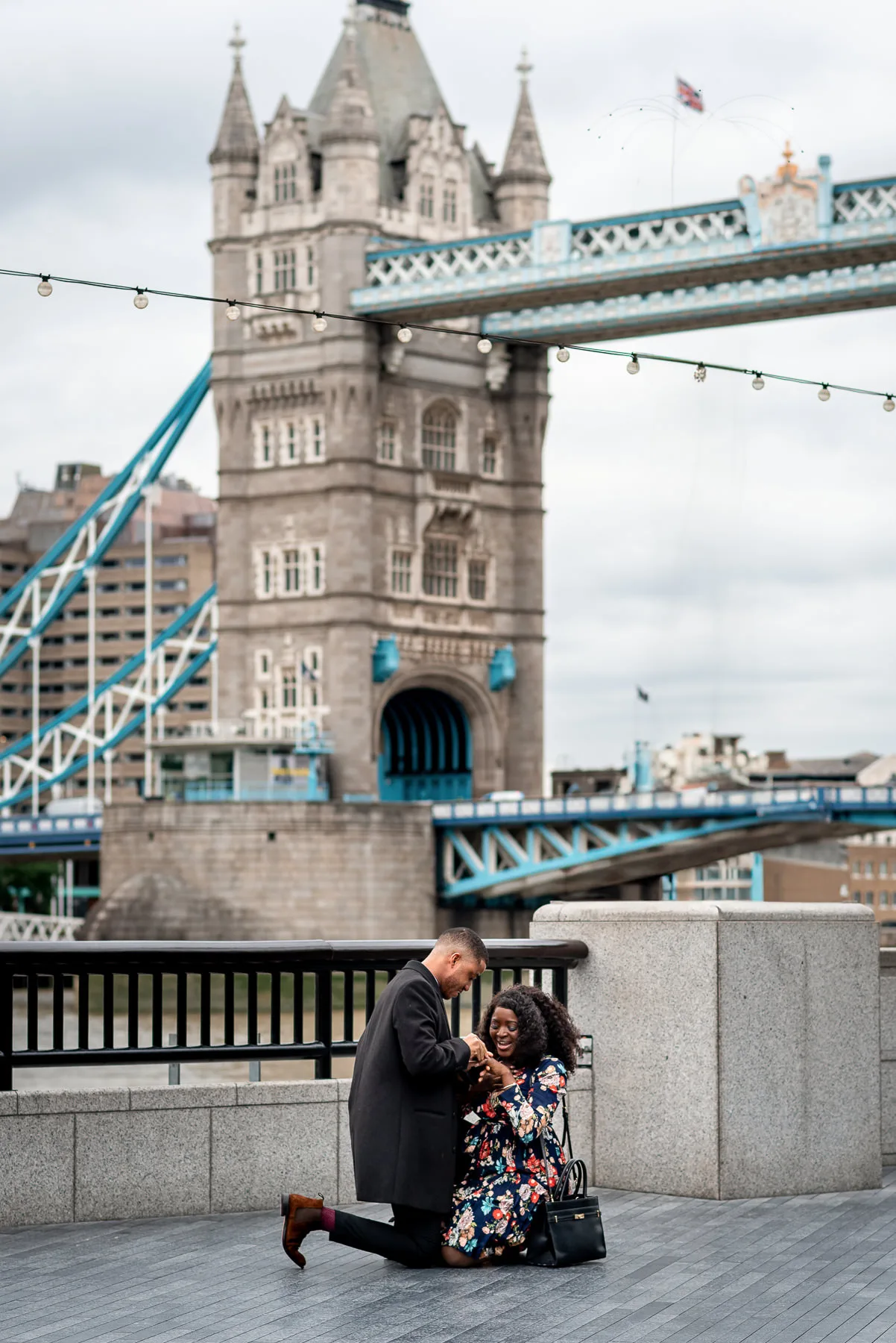 A man and woman are both on their knees in front of Tower Bridge after he proposes to her in London