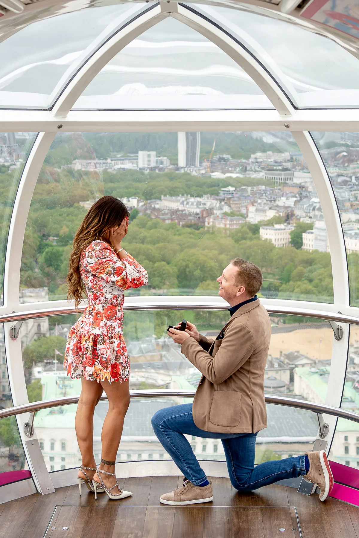 A man is on one knee proposing to his girlfriend on the London Eye as she covers her face with her hands in surprise. She said yes.