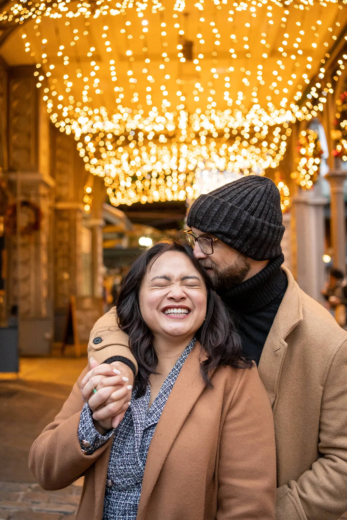 A man hugs and kisses his new fiancé with the Christmas lights of Borough Market behind them