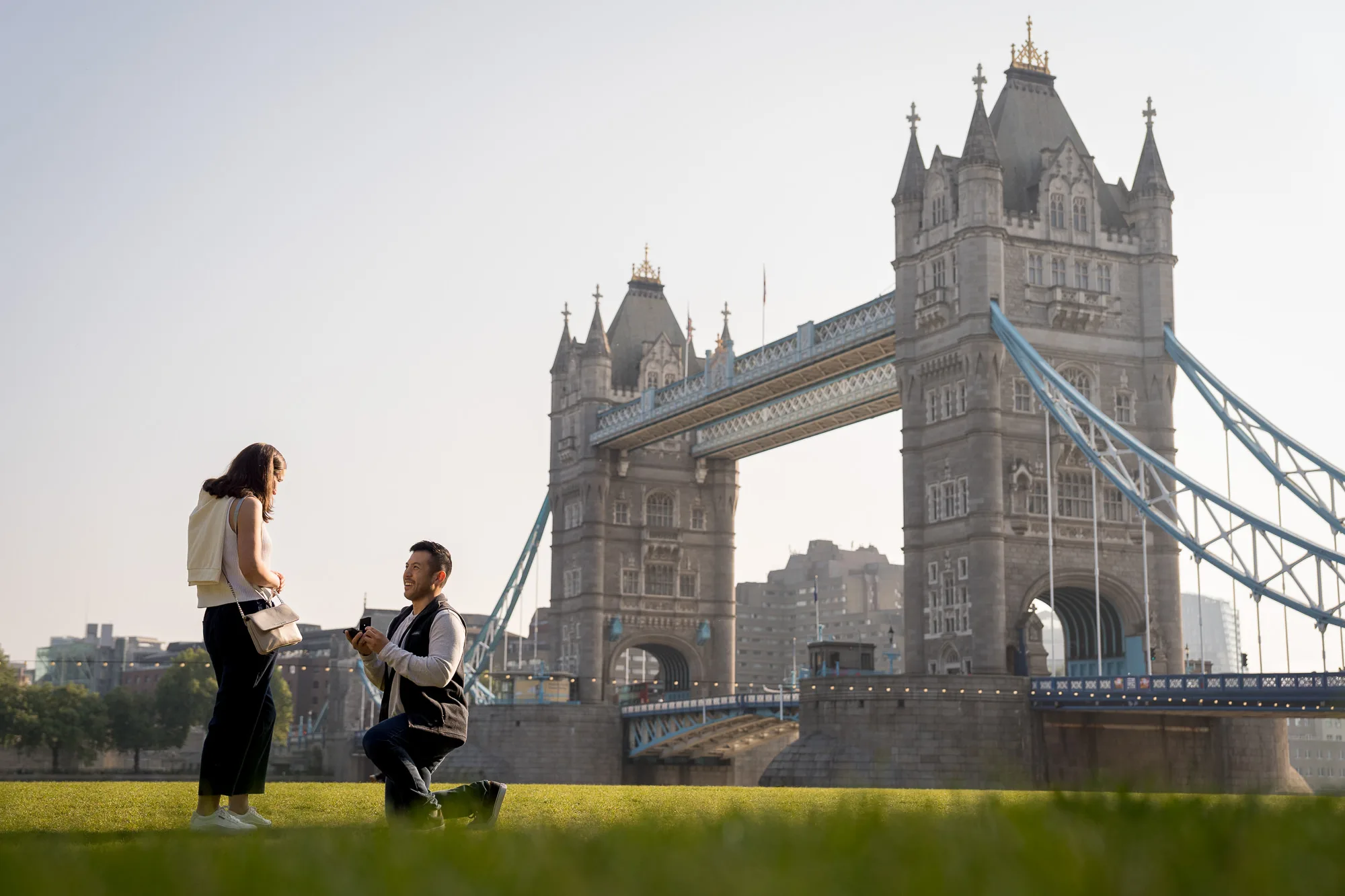 A man on one knee proposing to his girlfriend in front of Tower Bridge on a summer morning in London